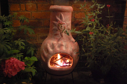 A nighttime shot of a clay chiminea with a blazing fire in a garden. A brick wall is in the background with hydrangea and valerian
